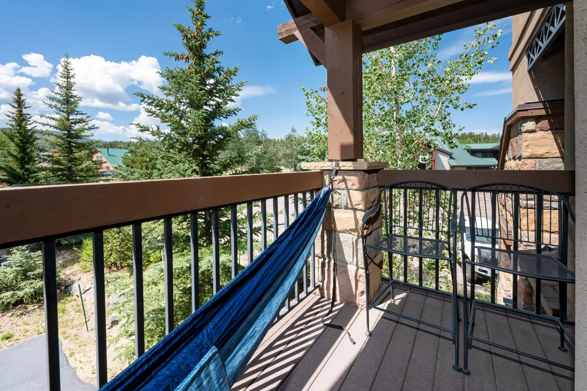 This image presents a tranquil balcony scene featuring a hammock gently swaying above a wooden deck, framed by a sturdy railing and surrounded by leafy trees. The sunlight filters through the branches, evoking the pleasure of relaxing outdoors, reading, or simply soaking in the fresh mountain air.