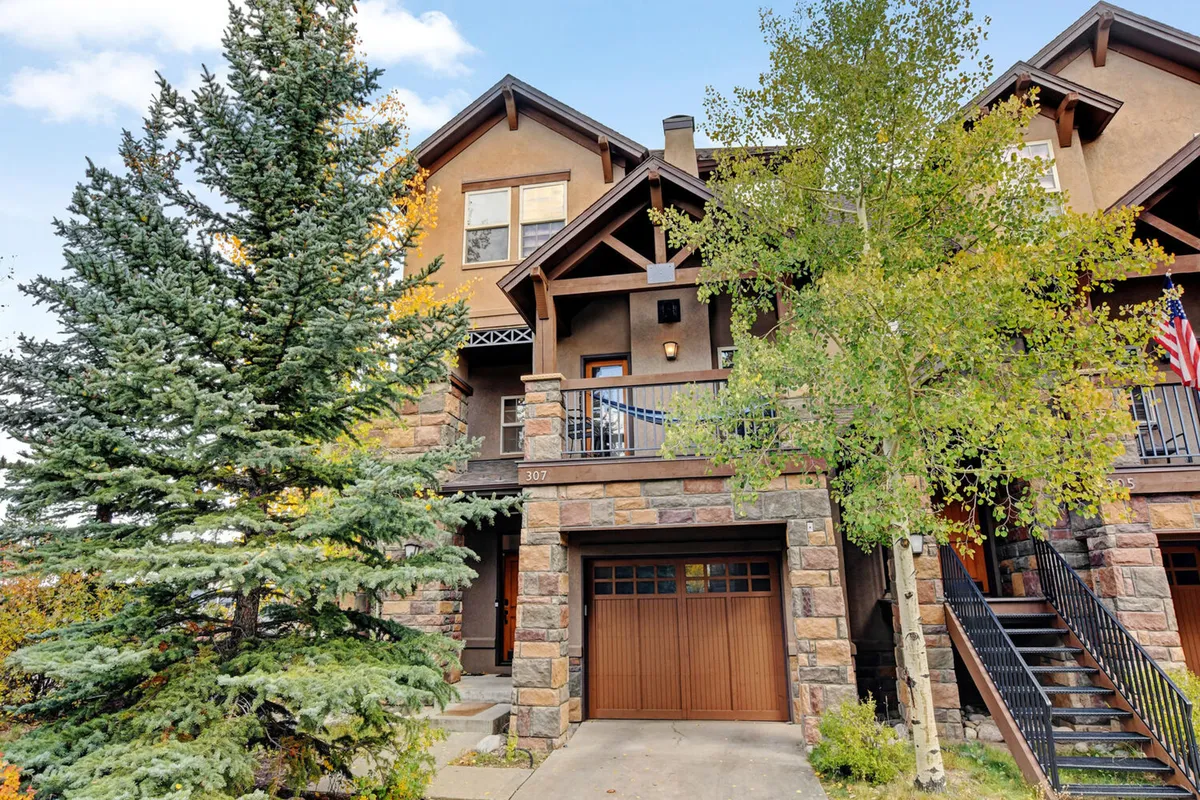 The image showcases the home's impressive stone façade and elegant wooden balcony, framed by decorative eaves and lush mountain greenery. The scene is inviting, suggesting opportunities for relaxing on the balcony, hosting gatherings, and enjoying the scenic natural surroundings.