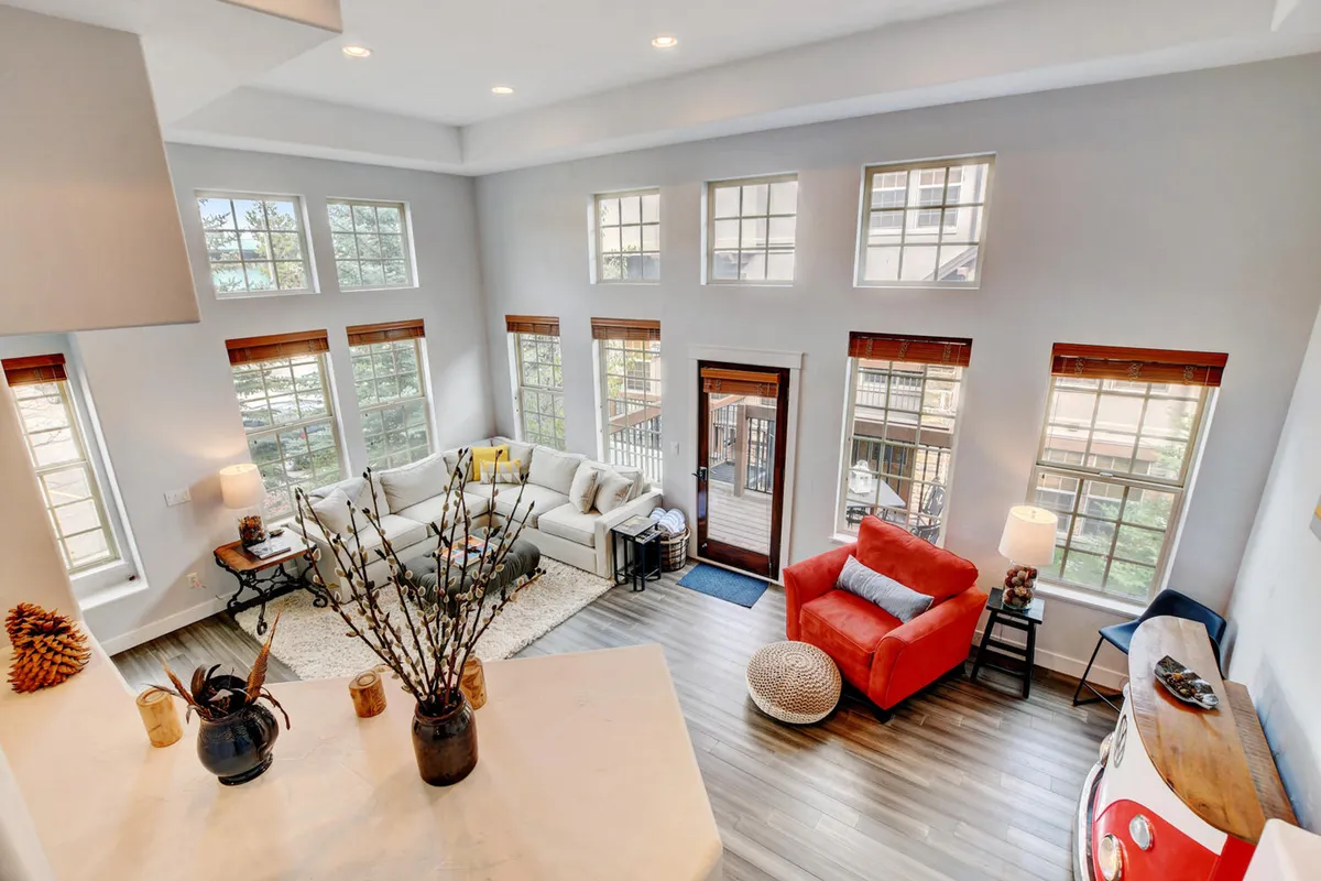 A bright and spacious living area with a large sectional sofa, red accent chair, stylish coffee table, and expansive windows offering beautiful mountain views. The setting conveys a sense of openness and connection to the Rockies, perfect for relaxing, socializing, or enjoying board games and movies.