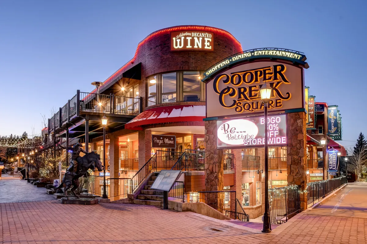 A bustling downtown scene featuring the Cooper Creek Square sign, a wine shop sign, outdoor seating, and a whimsical statue in the foreground. The area is lively, with people enjoying shopping, dining, and the energetic atmosphere, all set against a backdrop of charming storefronts.
