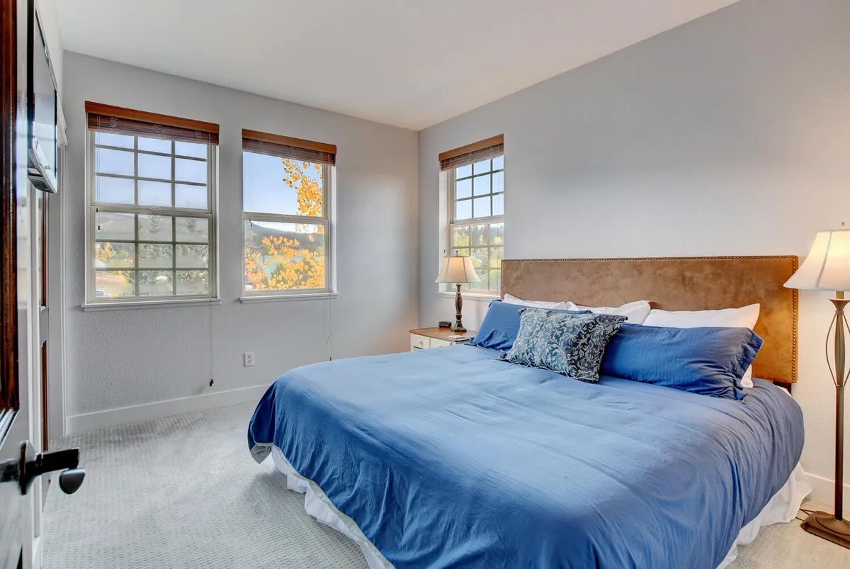 A serene bedroom highlighted by a king-sized bed with blue bedding, two windows streaming in natural light, and a nightstand with a lamp. The room feels calm and cozy, inviting relaxation and rest.