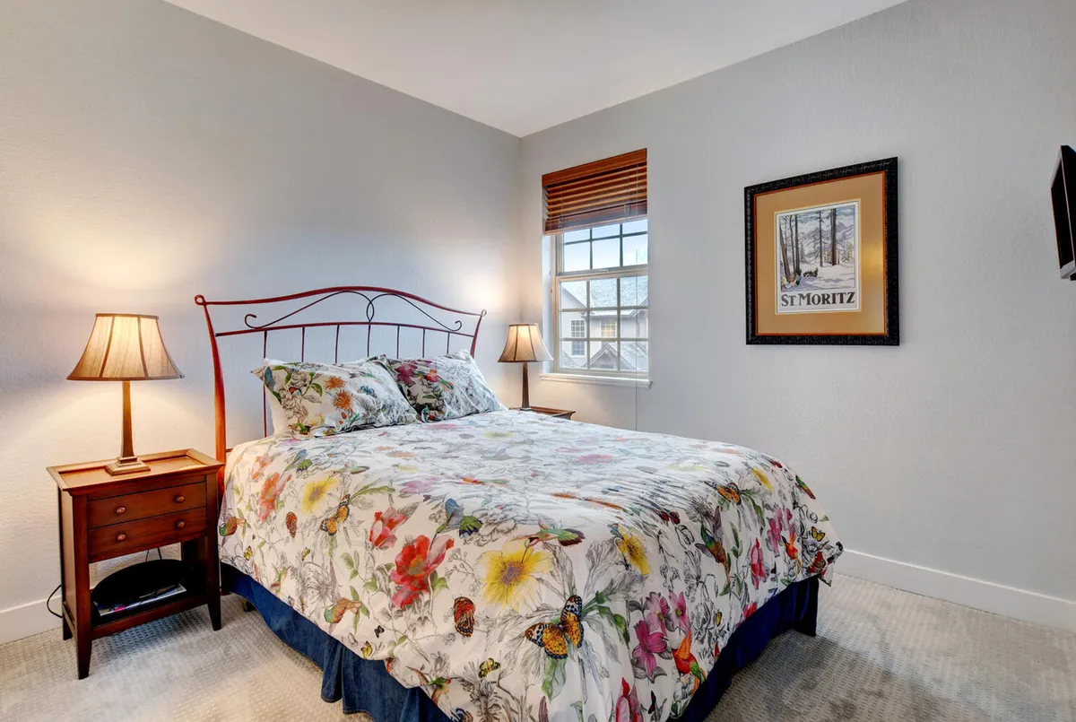 A light-filled bedroom with flower-patterned bedding on an iron bed frame, a nightstand with lamp, and a window with blinds. The room exudes a tranquil, inviting atmosphere perfect for morning coffee or a quiet read.