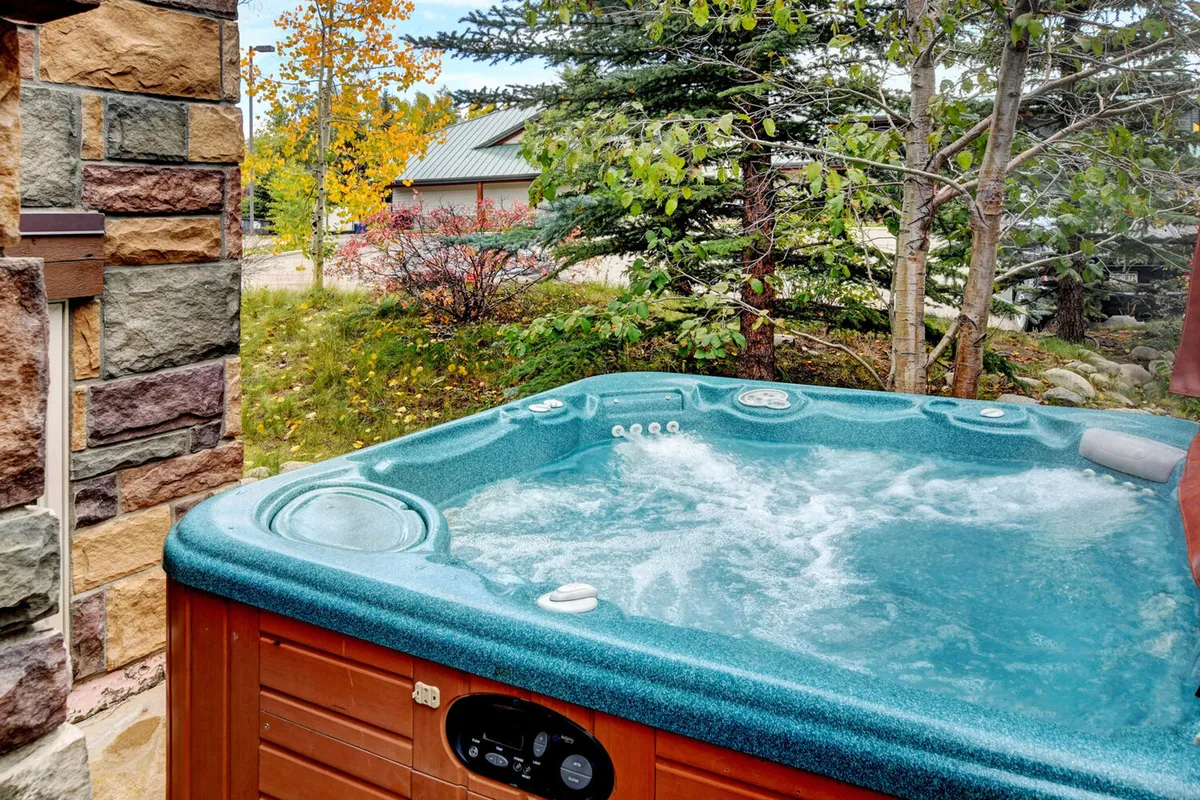 An exterior view focused on a bubbling private hot tub set against natural stone walls and surrounded by lush greenery. The scene suggests a space for relaxation and soaking while enjoying the peace of the outdoors.