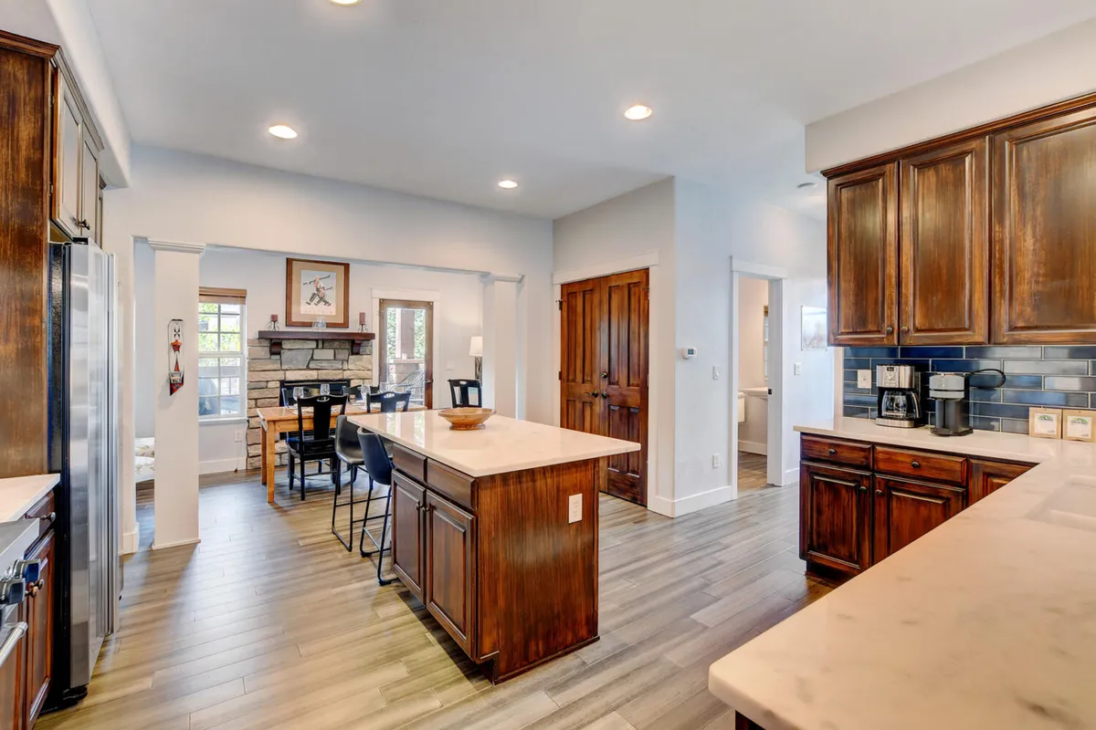 An open-concept kitchen and dining area with a generous island countertop, wooden cabinetry, a dining table adjacent to a cozy fireplace, and natural light streaming in. The space feels warm, communal, and inviting.