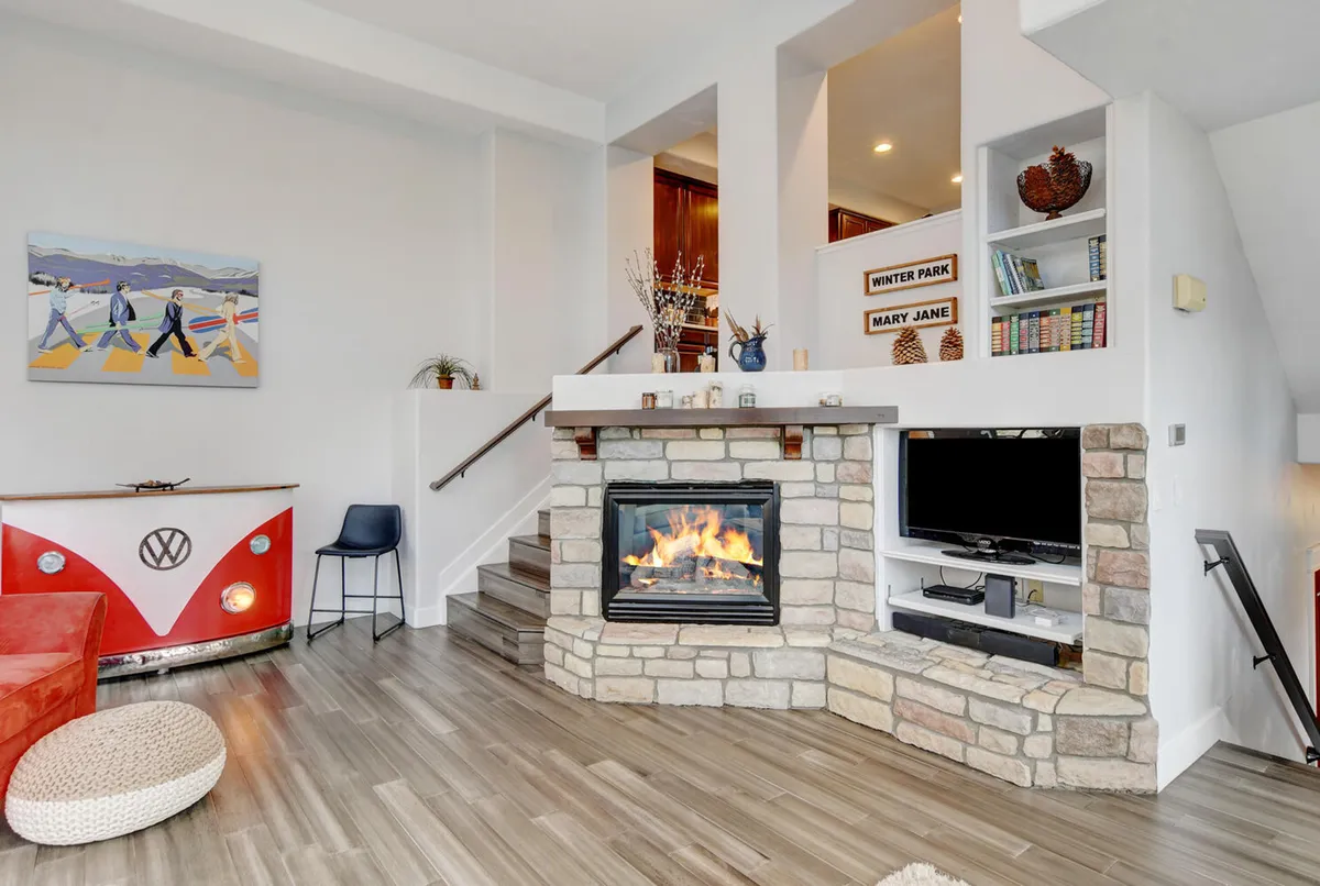 A cozy, inviting living area featuring a prominent stone gas fireplace, comfortable seating, a VW bus decorative element that echoes the custom bar theme, and large windows that let natural light flood in. The space exudes warmth and sociability, ideal for gathering and relaxing with friends or family.