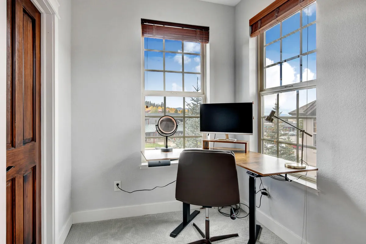 A dedicated workspace featuring a desk with monitor, comfortable chair, and broad windows flooding the room with natural light. The scene evokes focus and relaxation with views of the outdoors.